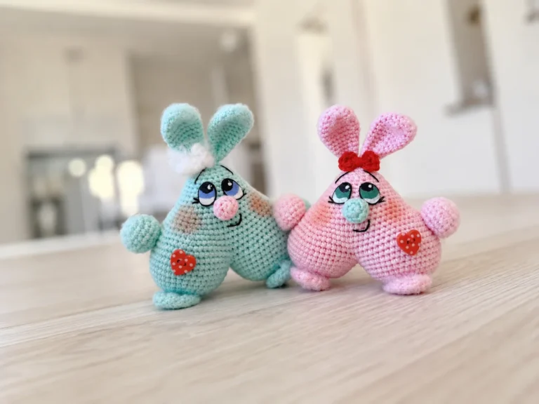 A pastel pink crochet bunny couple with heart-shaped bodies, painted eyes, and a red bow, sitting side by side.