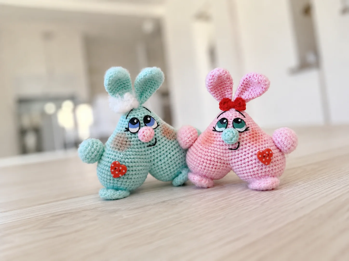 A pastel pink crochet bunny couple with heart-shaped bodies, painted eyes, and a red bow, sitting side by side.