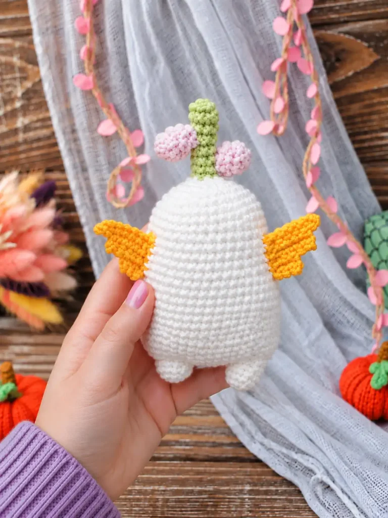 Hand holding the back of a white bulb amigurumi pattern toy showing yellow wings and green stem against rustic wood.