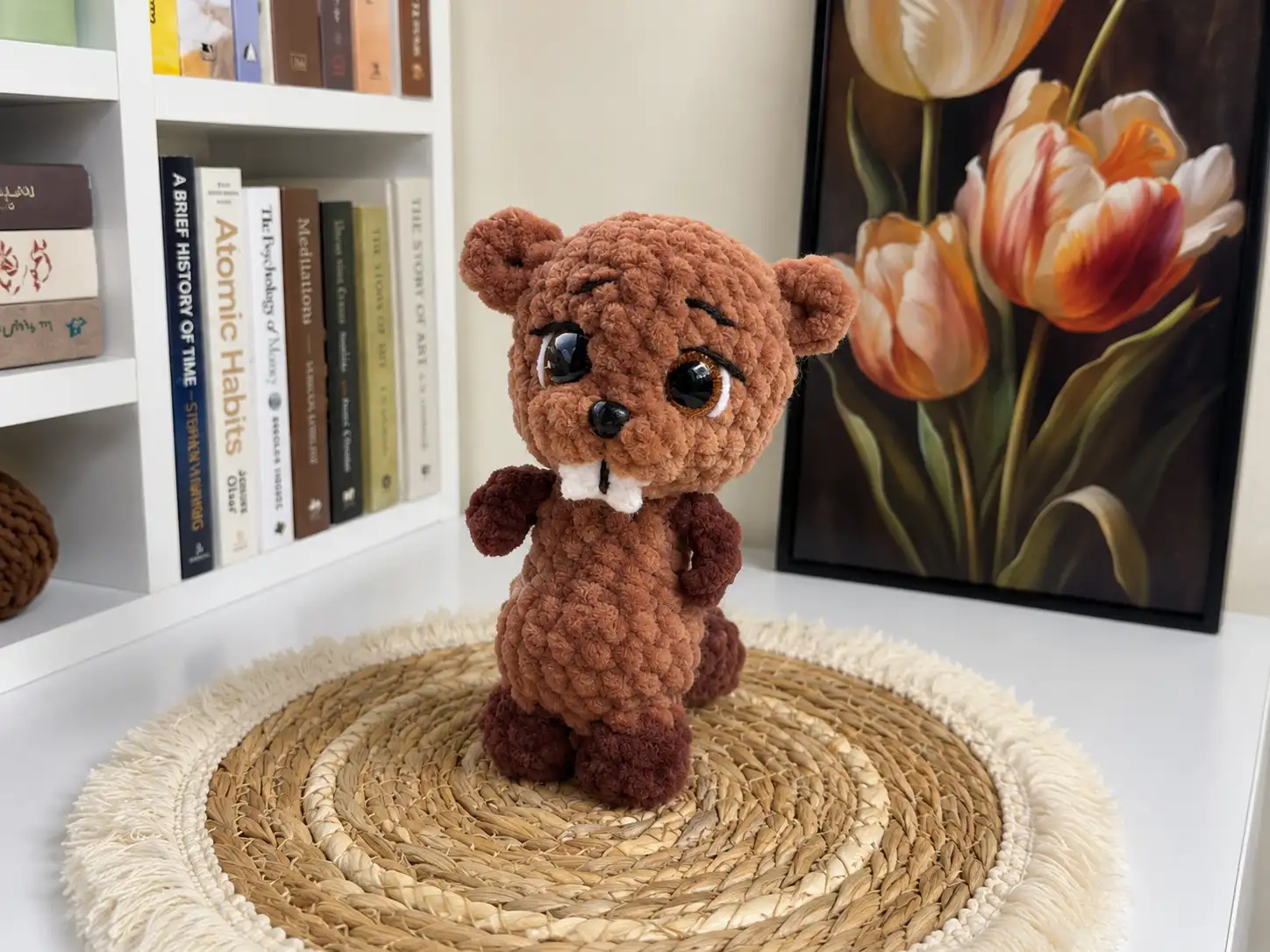 Plush brown beaver amigurumi toy with white front teeth, standing on a woven straw mat in a bright room.