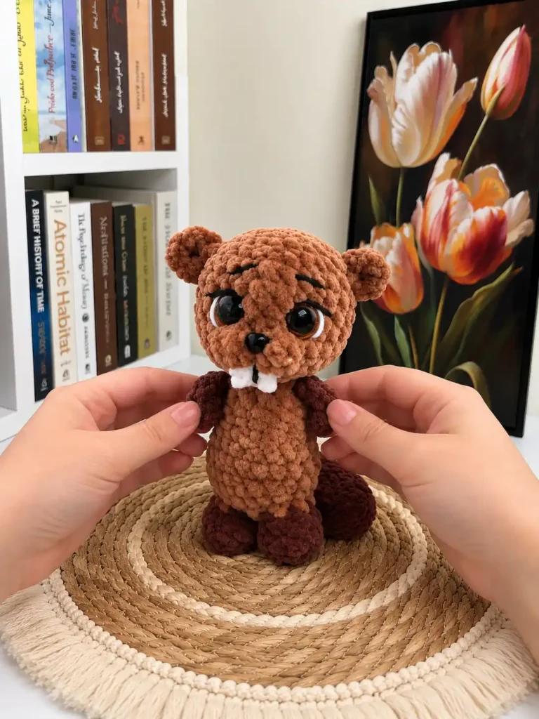 Two hands holding the plush brown beaver amigurumi toy with white teeth over a round woven straw mat.