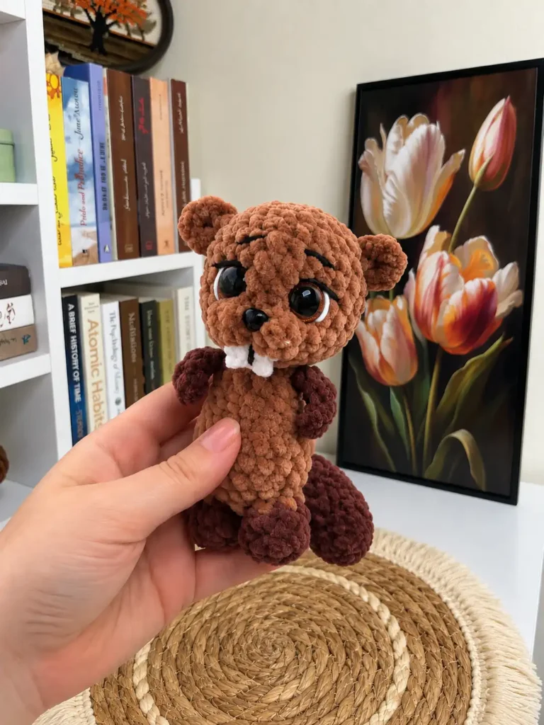 A hand holding up the cute brown plush beaver amigurumi toy with shiny safety eyes and white front teeth.