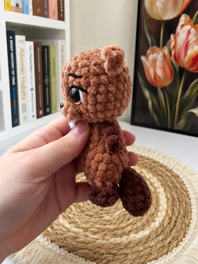 Side profile of the plush brown beaver amigurumi toy showing its rounded head, small ear, and textured tail.