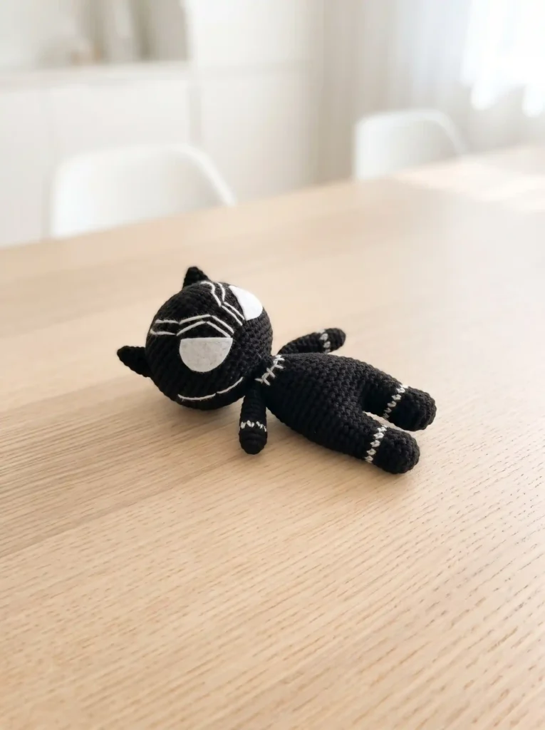 Black panther amigurumi crochet doll lying flat on a light natural wood dining table.