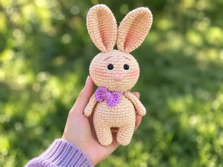 A hand holding a beige crochet bunny plush with long floppy ears and a purple bow against a bright background.