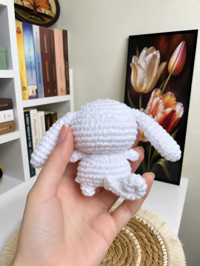Hand holding the back of a white Cinnamoroll crochet pattern toy, showing the twisted tail, with a bookshelf background.