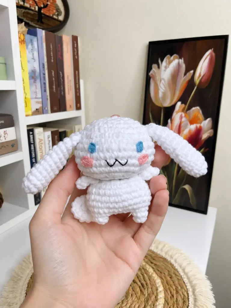 Hand holding a finished white toy from our Cinnamoroll crochet pattern, showing blue eyes and pink cheeks by a bookshelf.