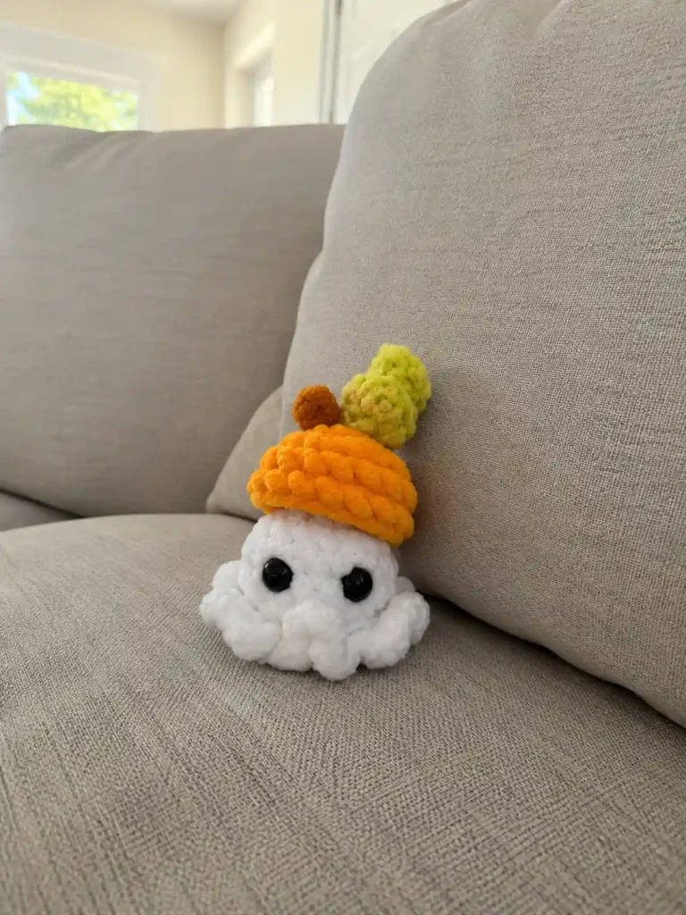 A white plush yarn toy from a ghost amigurumi pattern with an orange pumpkin hat sitting on a beige couch cushion.