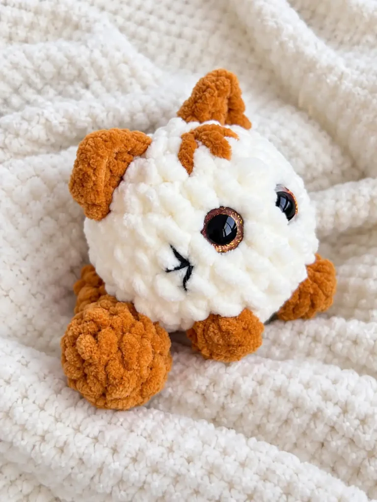 A soft white and brown plush kitten amigurumi pattern resting comfortably on a chunky white textured knit blanket.