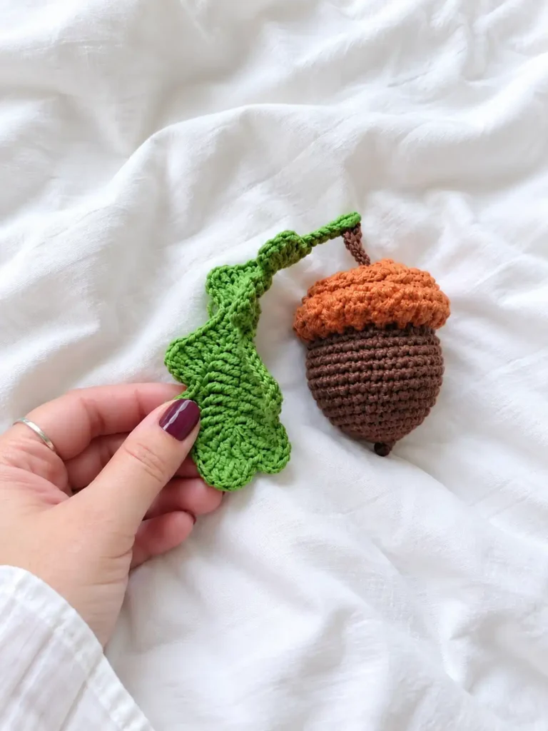 Hand holding the green leaf of a stuffed toy from a crochet acorn pattern resting on wrinkled white fabric.