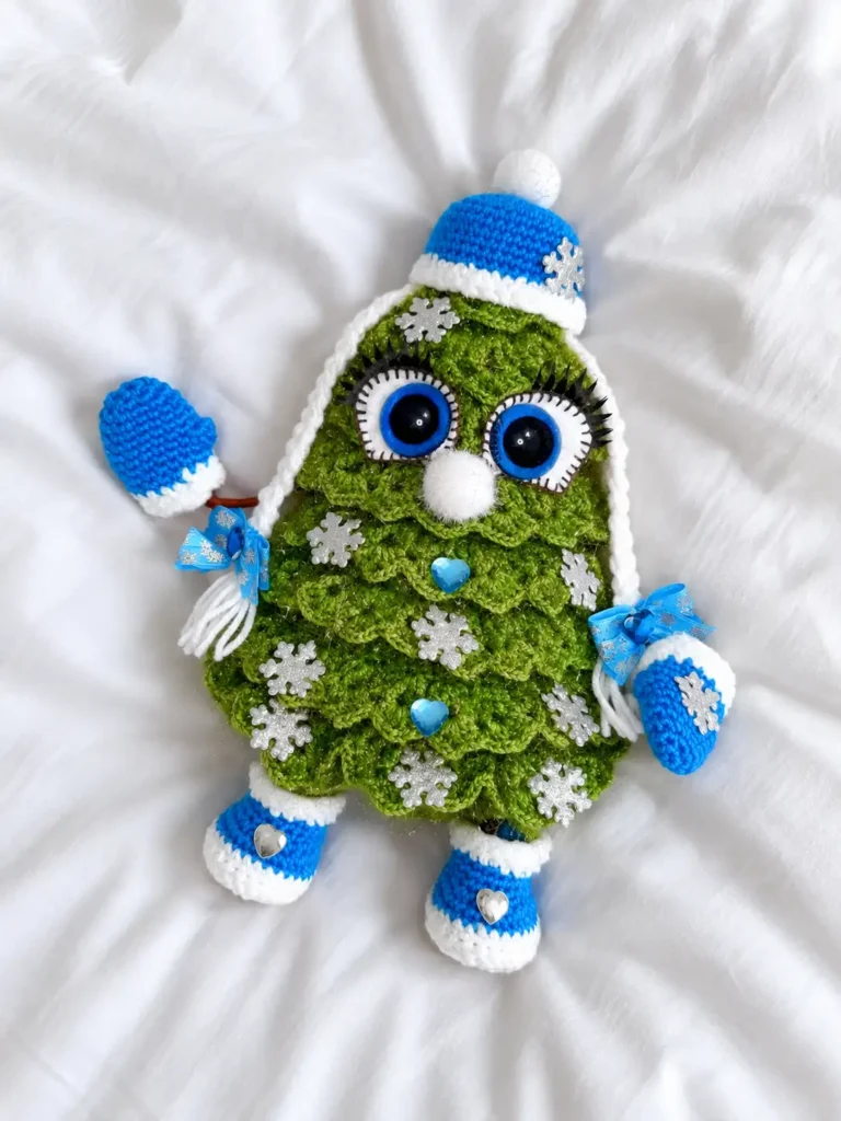 Plump green crochet Christmas tree toy with blue hat and mittens lying flat on a white knitted blanket.