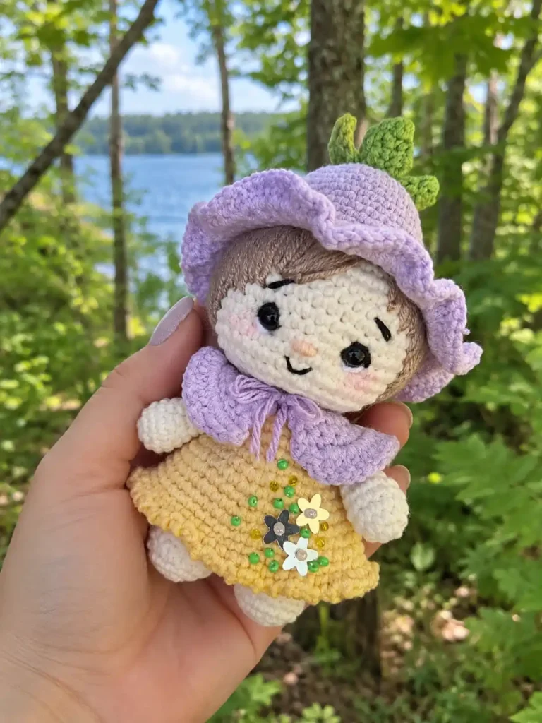 Hand holding a cotton yarn crochet gnome pattern doll with Alize Extra hair, purple hat, and yellow skirt by a lake.