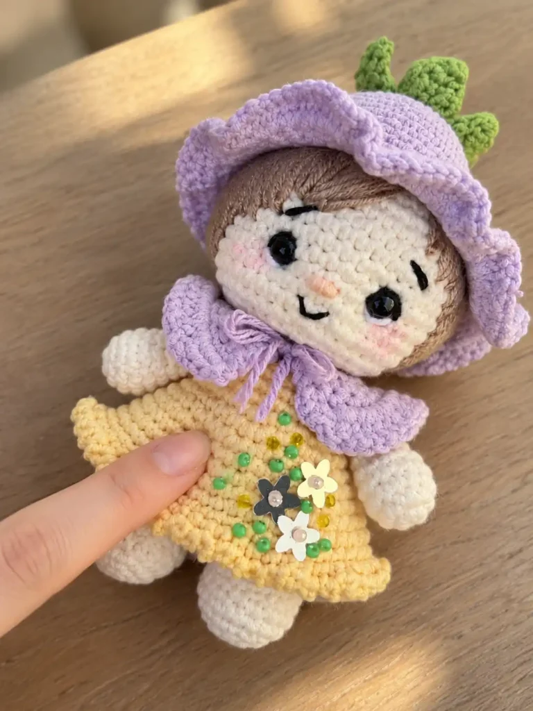 Finger touching the yellow cotton yarn skirt of a crochet gnome pattern doll resting flat on a light wooden table.