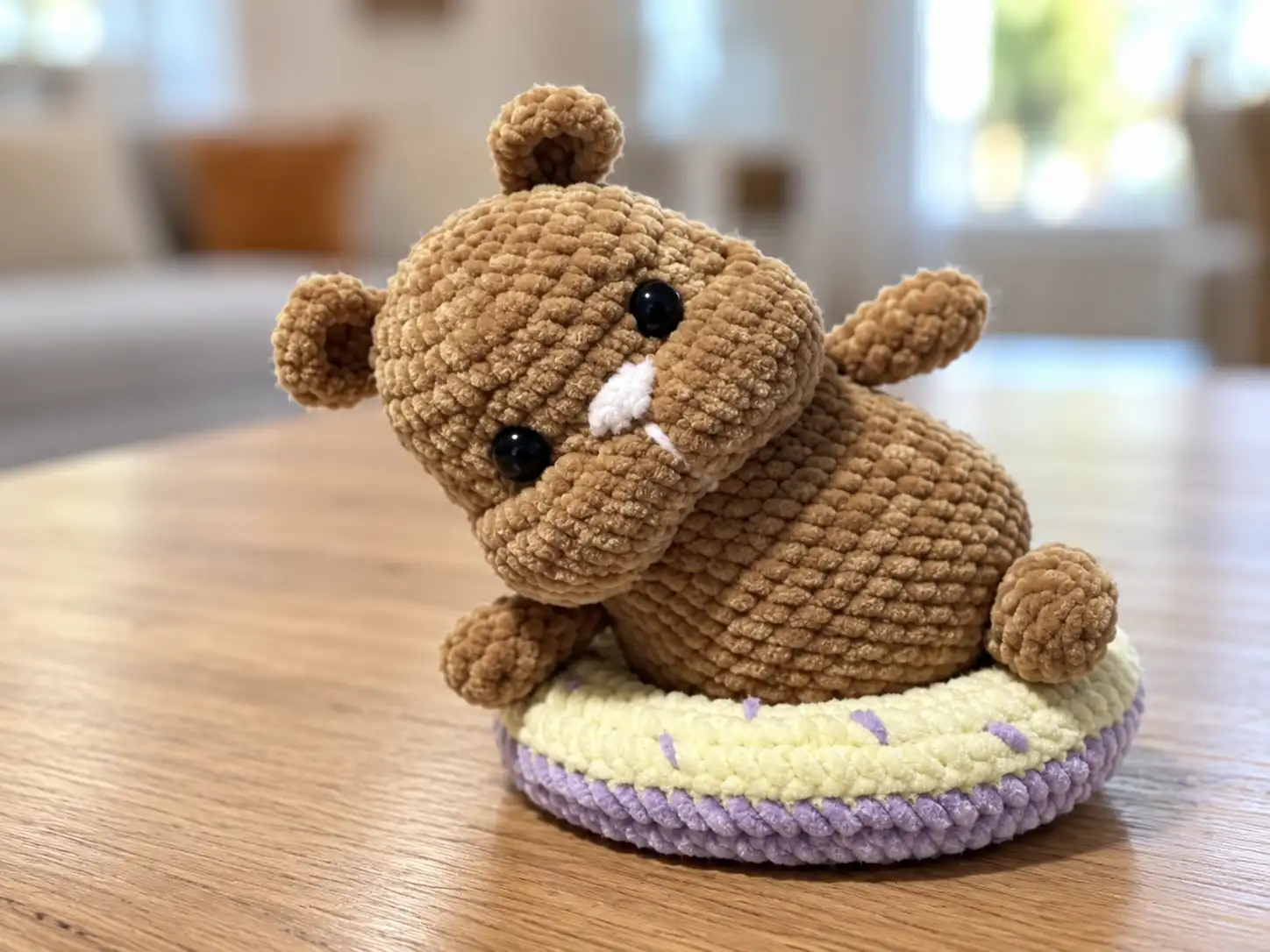 Brown chenille crochet hamster lying on a yellow and purple crochet donut on a white table.