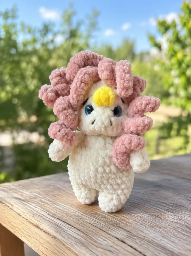 A white curly unicorn amigurumi pattern plush with pink hair standing on a wooden table outdoors with green trees behind it.
