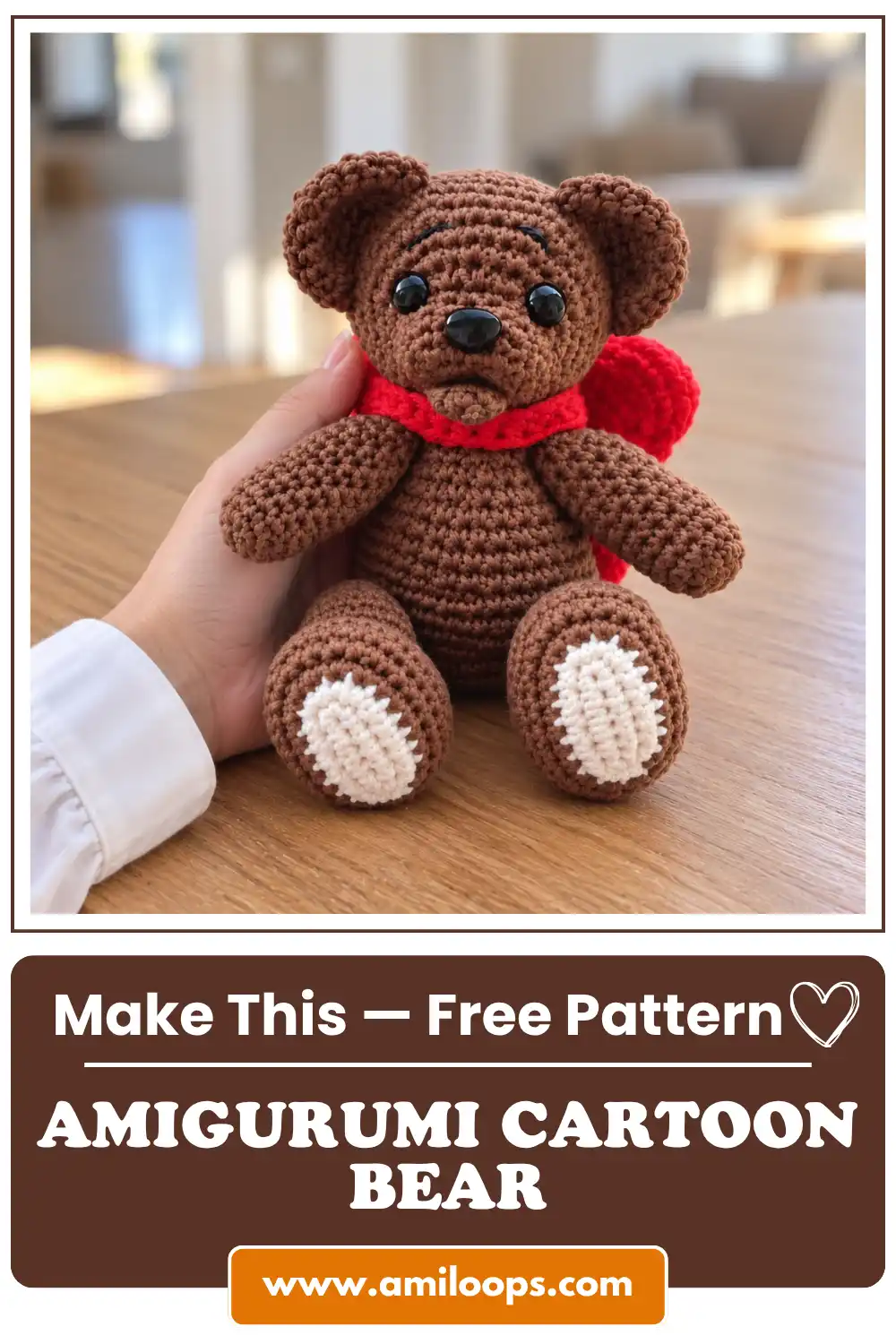 A cute brown amigurumi cartoon bear with a red scarf sitting on a wooden table, gently held by a hand in a bright room.