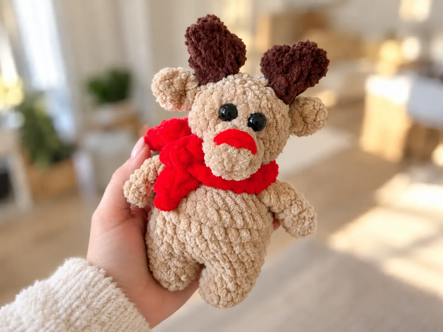 Hand holding a beige plush toy from a deer in a scarf crochet pattern with a red scarf against a blurred room background.