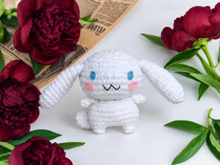 A white crocheted amigurumi Cinnamoroll toy with blue eyes, surrounded by maroon peonies and vintage paper.