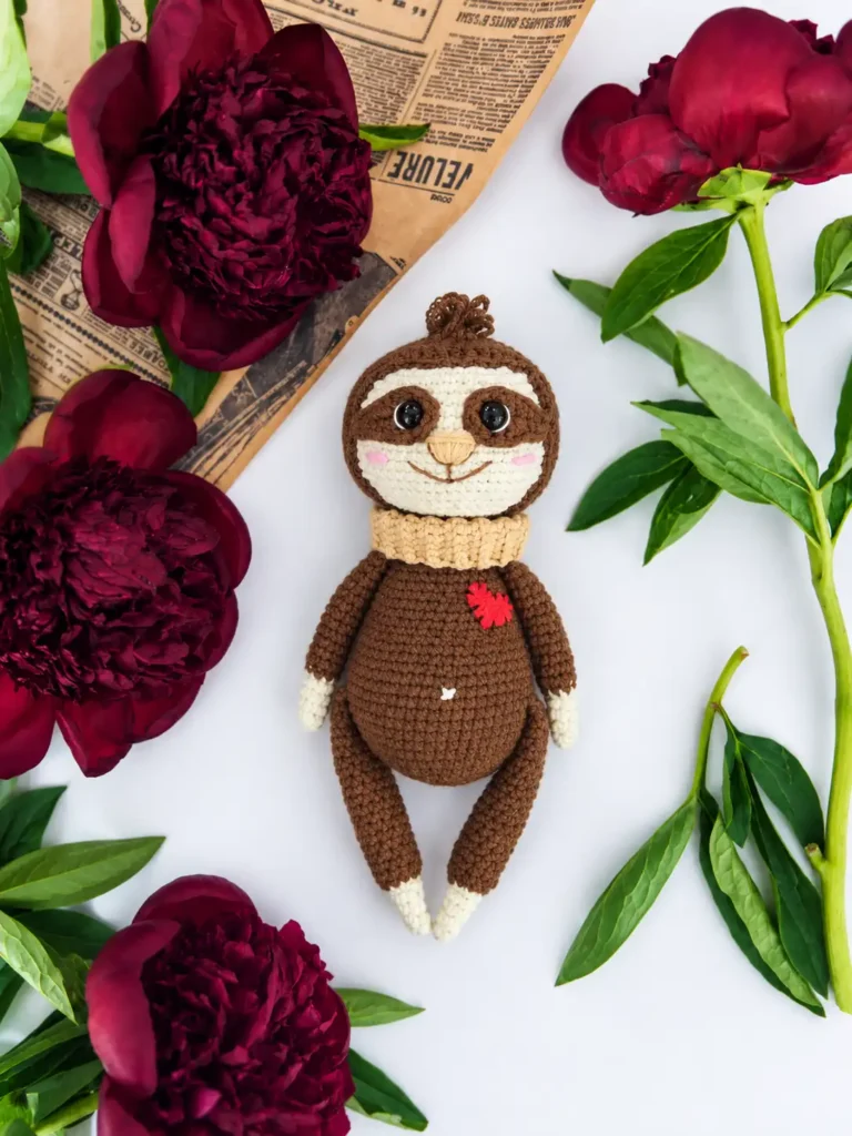 A completed amigurumi sloth toy from this free crochet sloth pattern, lying surrounded by maroon peonies and vintage newspaper on white linen.