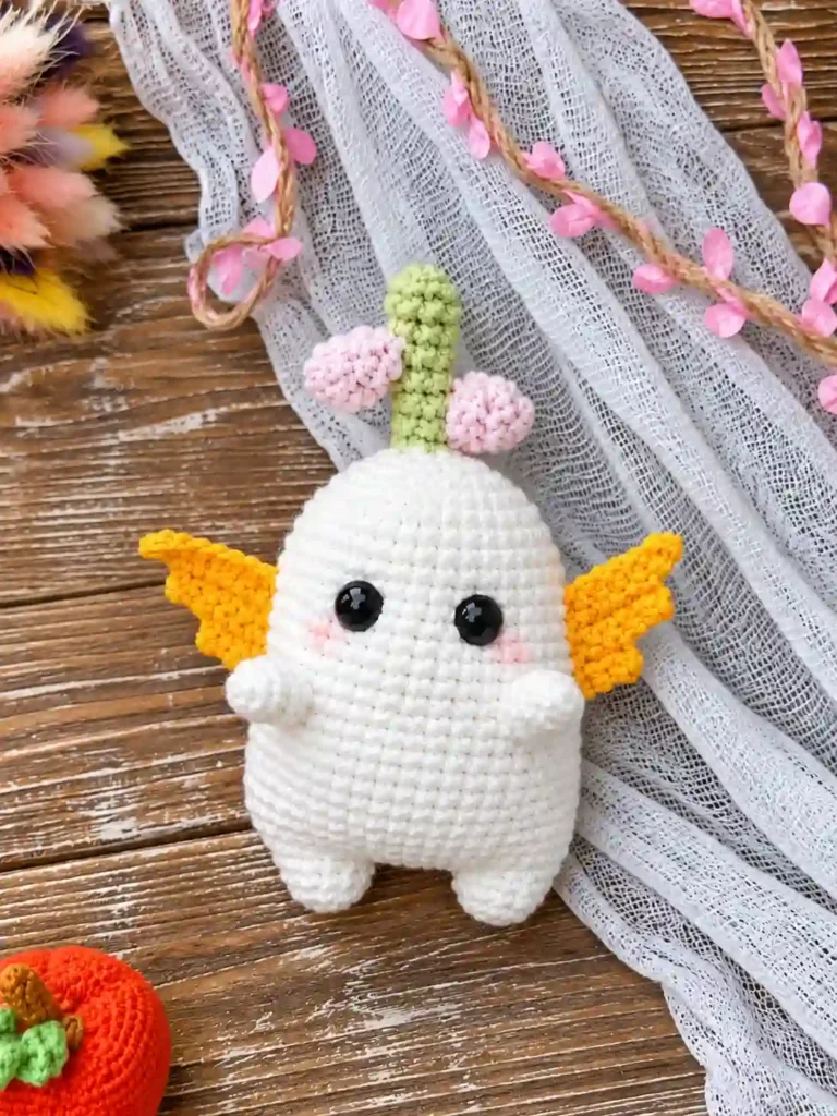 Front of white bulb amigurumi pattern toy with yellow wings on rustic wood with blue fabric and an orange crochet pumpkin.