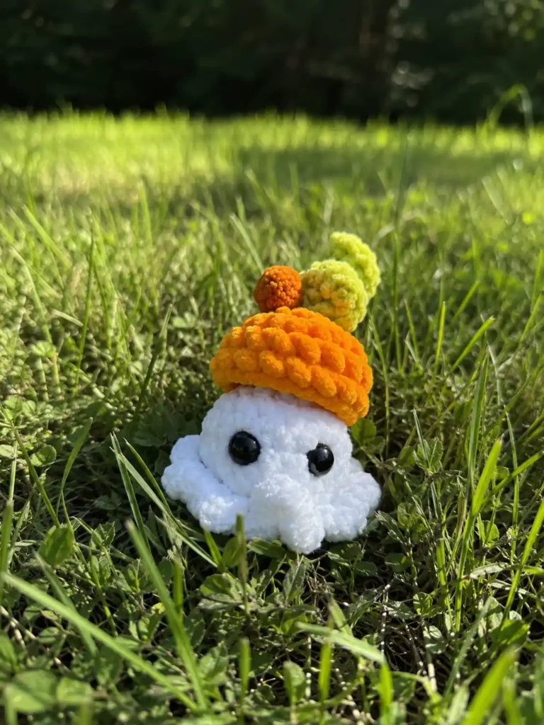A plush white toy made from a ghost amigurumi pattern, wearing an orange hat, sitting outdoors in short green grass.