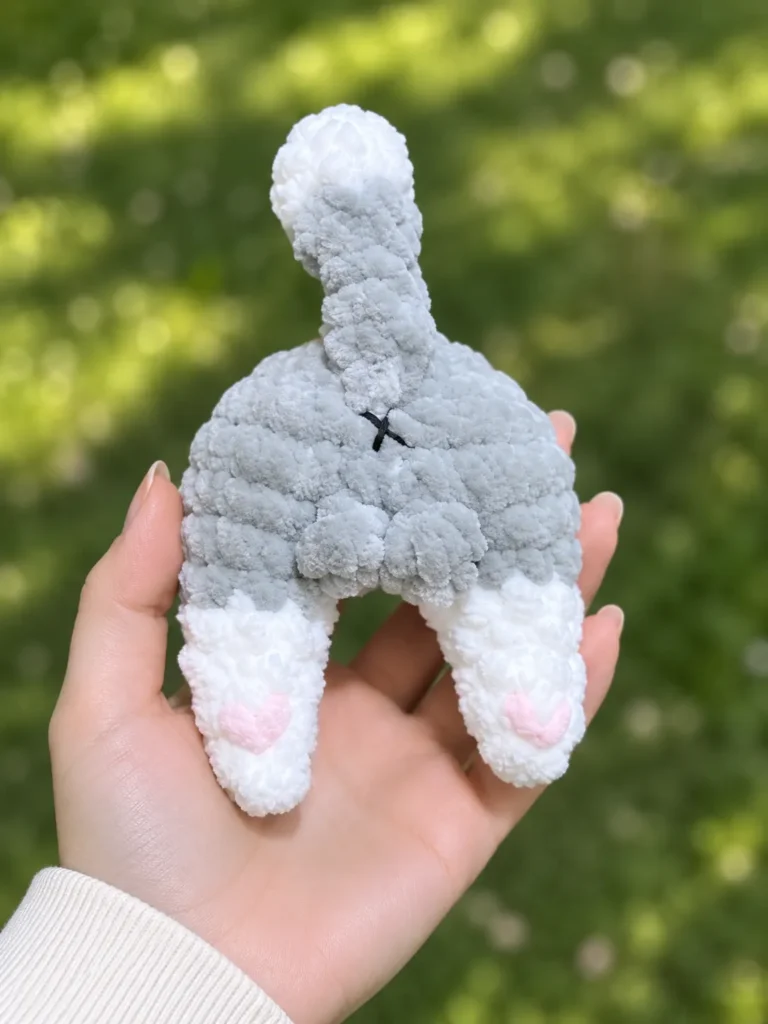 Hand holding a grey and white fluffy toy from a cat butt crochet pattern showing pink heart paws against a green background.