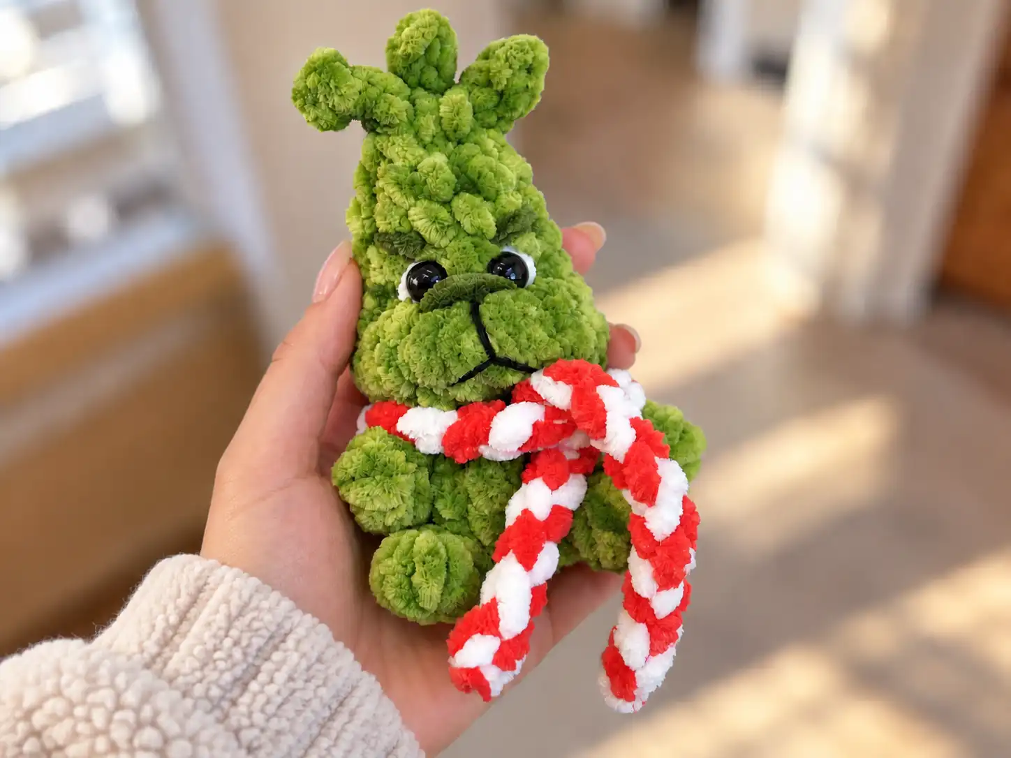 Hand holding a fluffy green grinch amigurumi crochet pattern toy with a red and white scarf in a bright room.
