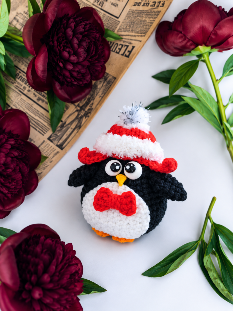 A holiday penguin amigurumi sporting a red earflap hat and bow tie, surrounded by dark red peonies and vintage newspaper.