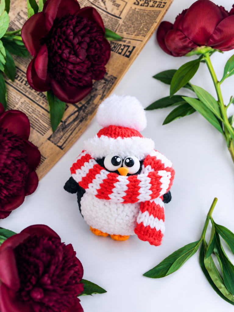 A holiday penguin amigurumi in a striped pom-pom hat and scarf, resting among red peonies and vintage newspaper on white.