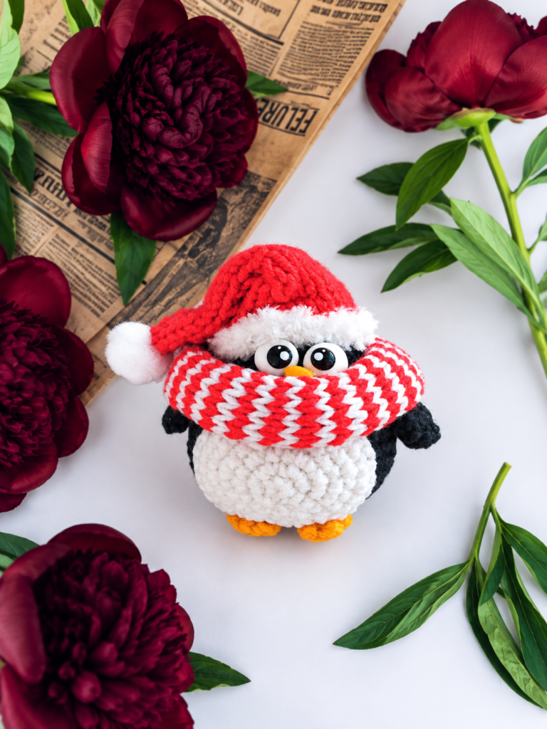 Top view of a holiday penguin amigurumi in a red stocking cap and striped snood, placed among red peonies and vintage paper.
