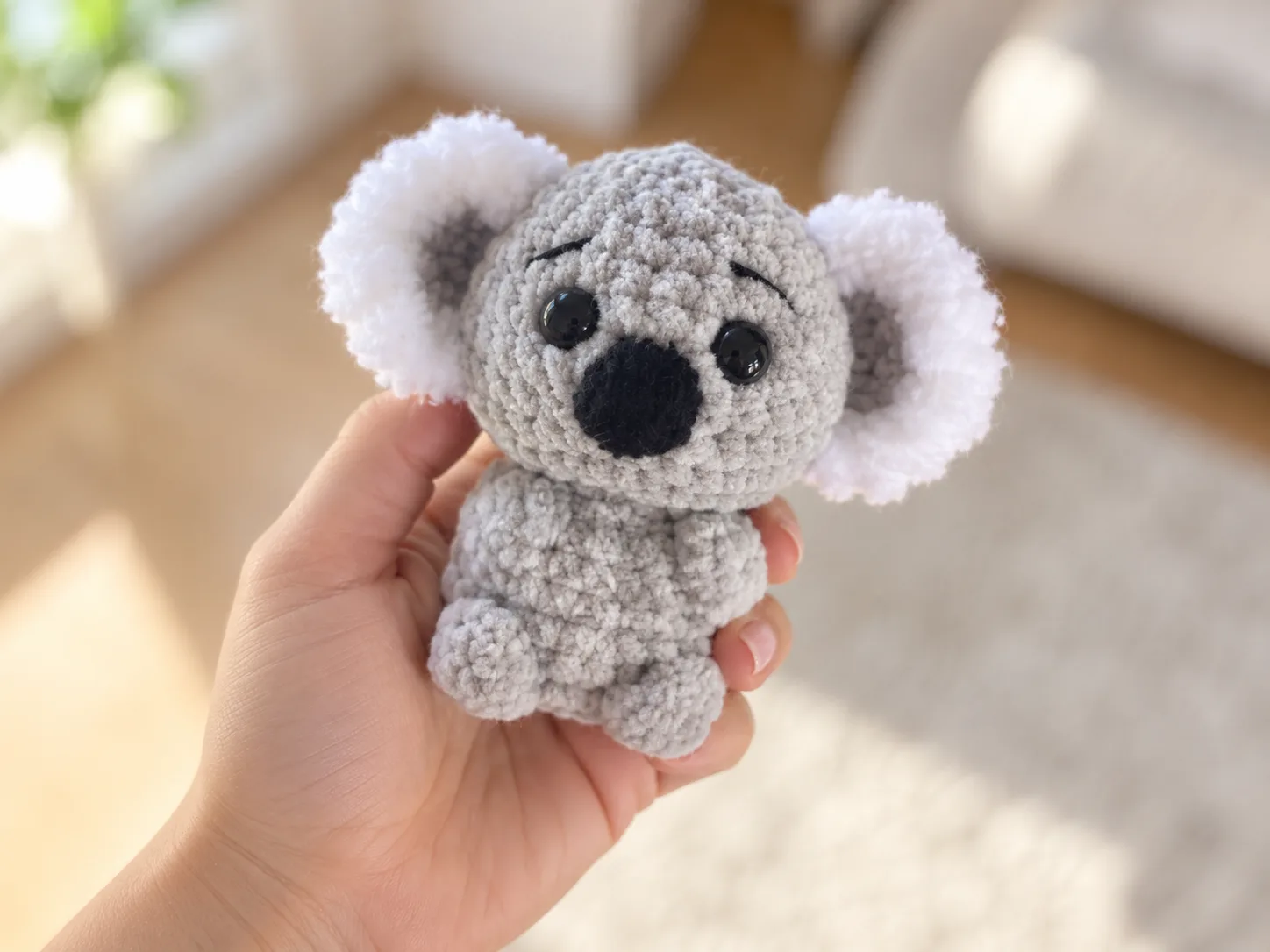 A gray crochet koala amigurumi with fluffy white ears and a black embroidered nose sitting on a woven mat.