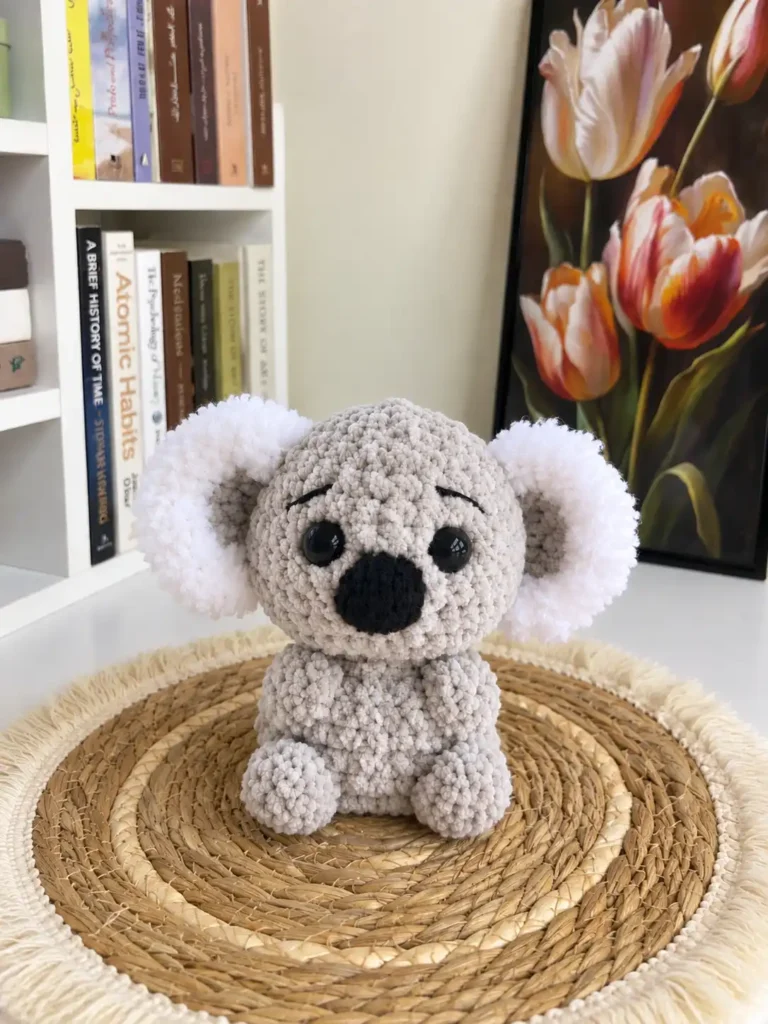 Plush grey koala amigurumi crochet pattern toy on a woven mat beside books and floral art.