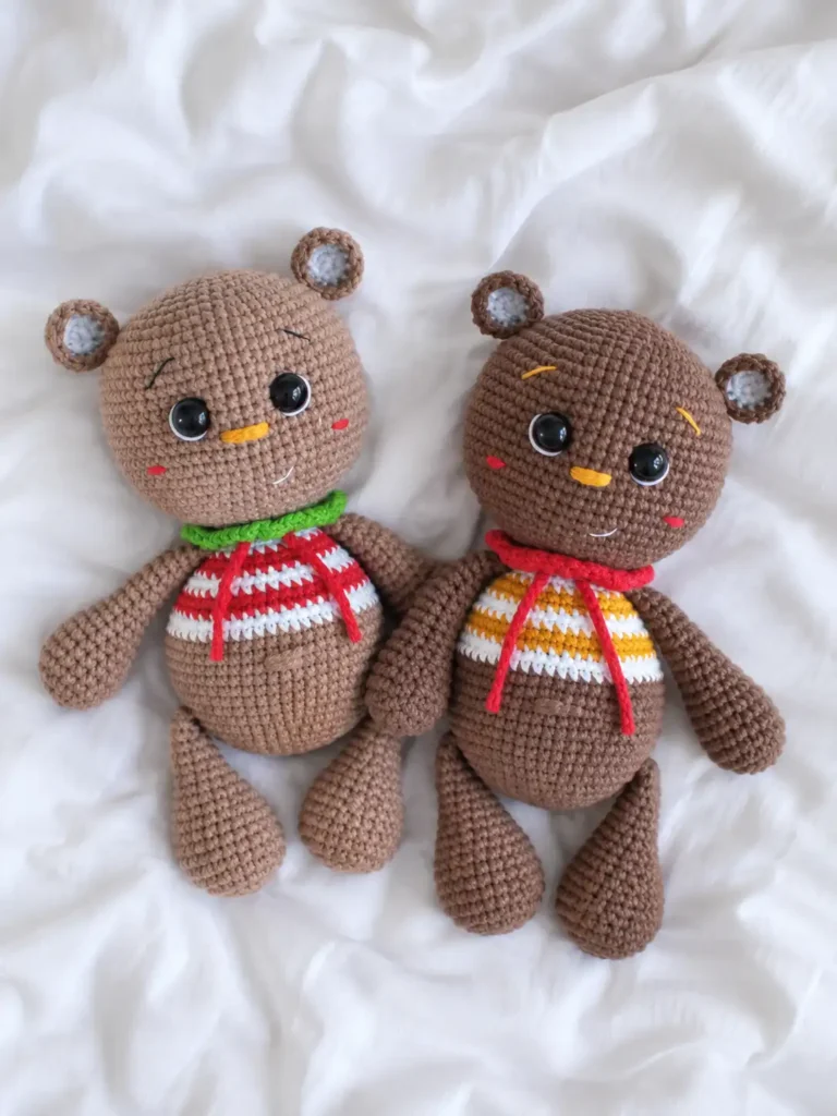 Two brown crocheted little bear in a sweater amigurumi toys laid flat on a wrinkled white fabric background.