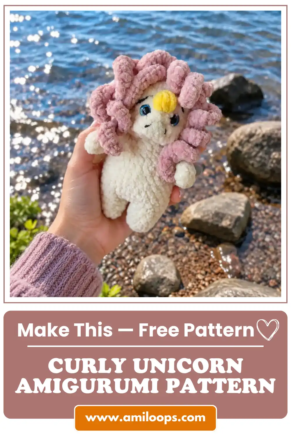 Hand holding a white curly unicorn amigurumi pattern toy with pink hair outdoors against a sparkling blue lake and rocks.