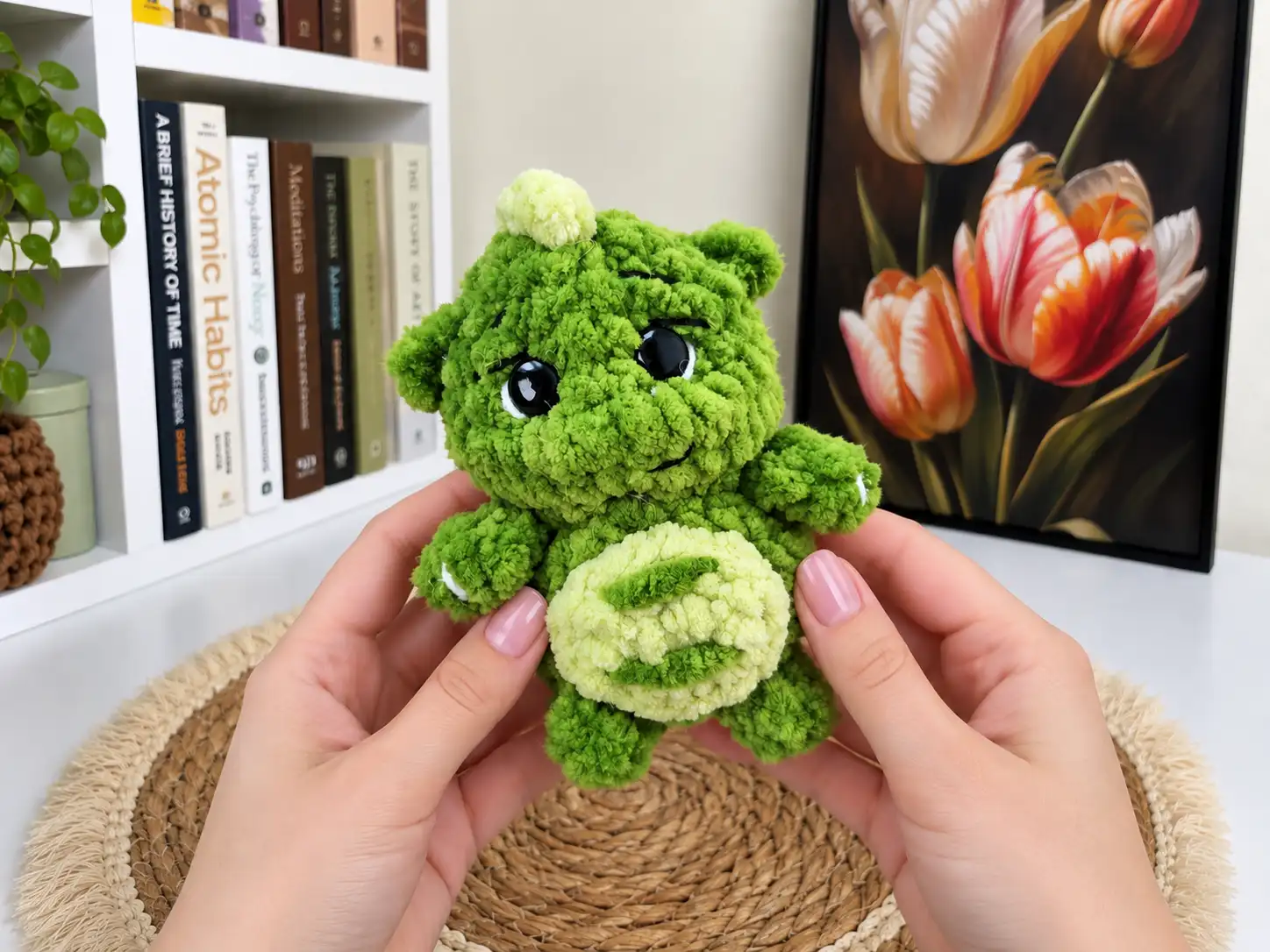 Hands holding a green plush mini dragon crochet pattern toy with large black eyes and a light green belly.