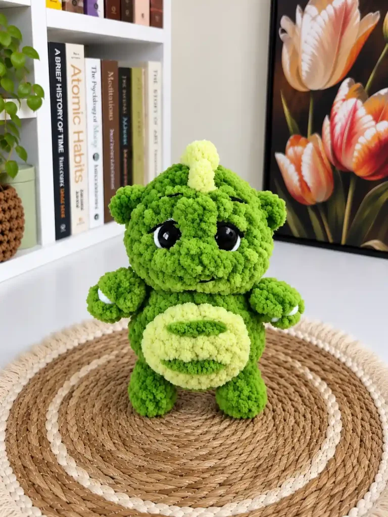 A green plush mini dragon crochet pattern toy standing upright on a round woven mat in front of a bookshelf and a tulip painting.
