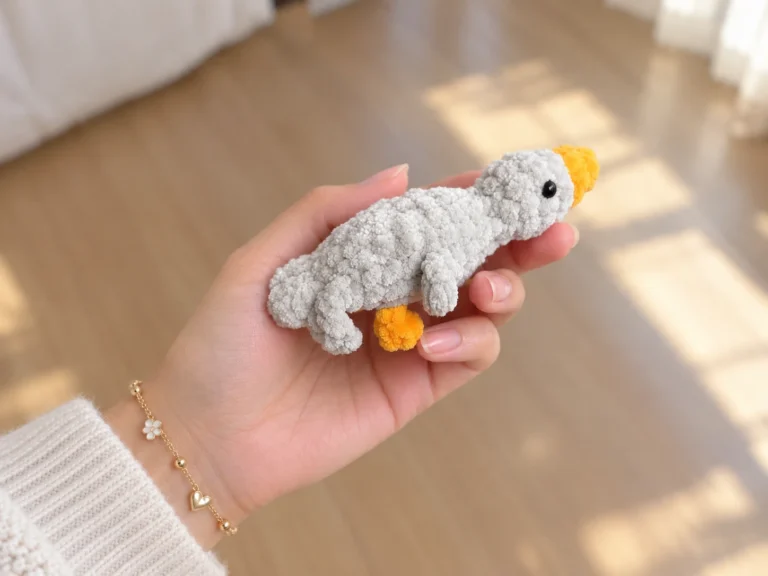 Hand holding a grey fuzzy mini gosling crochet toy with a yellow beak against a wooden floor