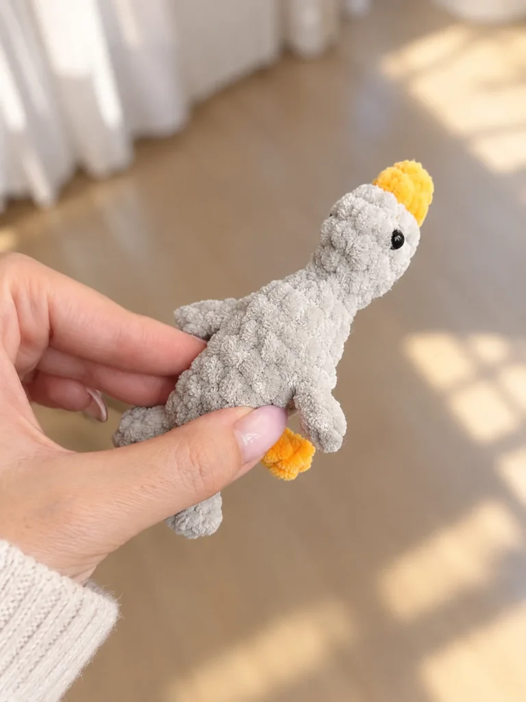 Hand holding a grey and yellow plush mini gosling crochet toy vertically in bright indoor light