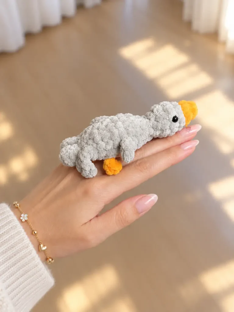 Hand holding a grey fuzzy mini gosling crochet toy horizontally over a sunlit wooden floor