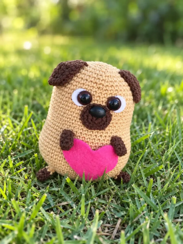Mini pug crochet pattern toy with brown ears and a pink felt heart, sitting outdoors in vibrant green grass.