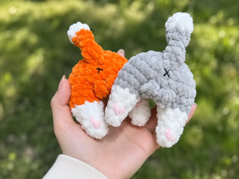 Hand holding orange and grey fluffy yarn toys from a cat butt crochet pattern against a blurred green outdoor background.