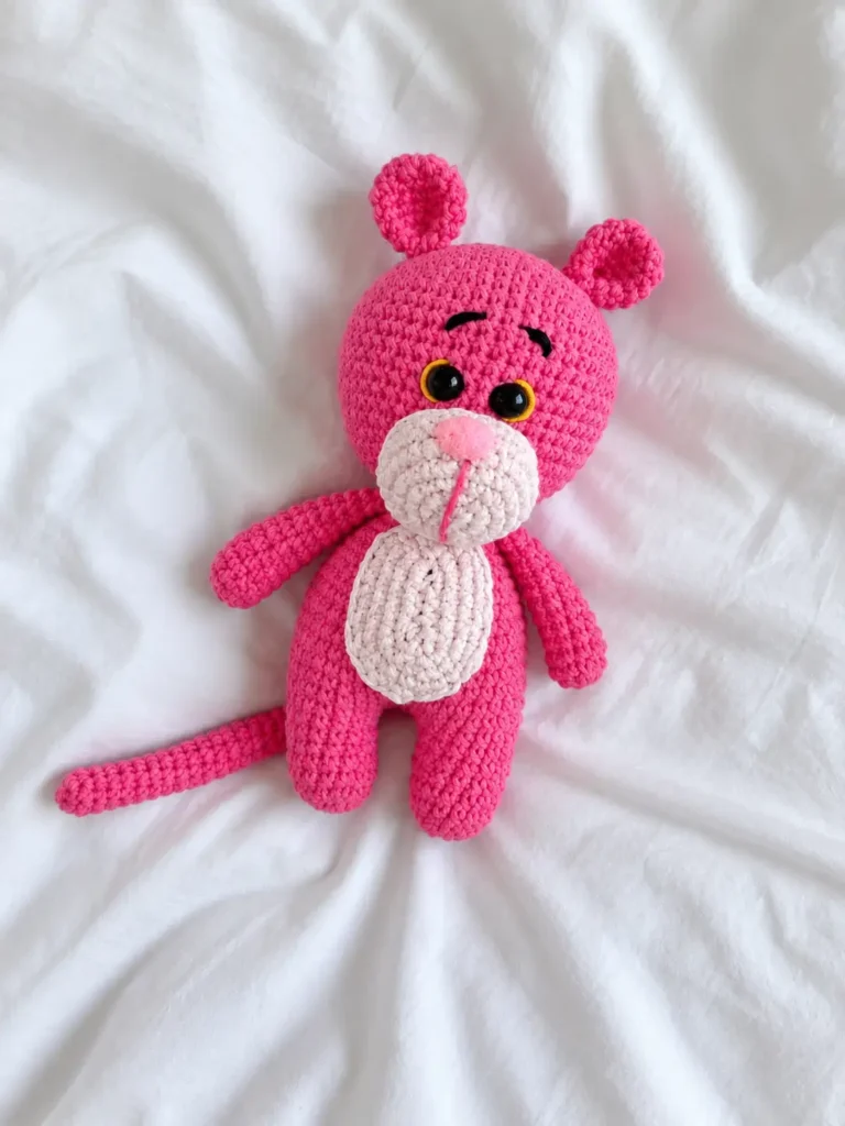 A completed bright pink yarn toy from our Pink Panther crochet pattern laying flat on a white wrinkled fabric surface.