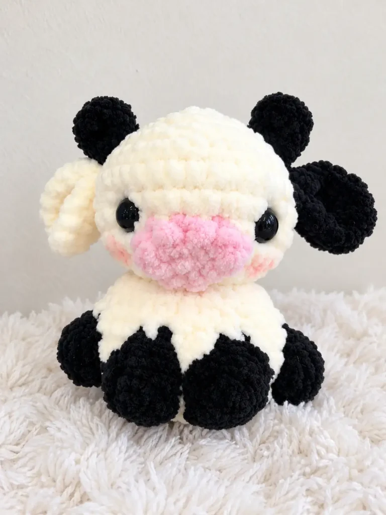 Cream and black plush cow amigurumi toy with a pink snout and safety eyes on a white background.