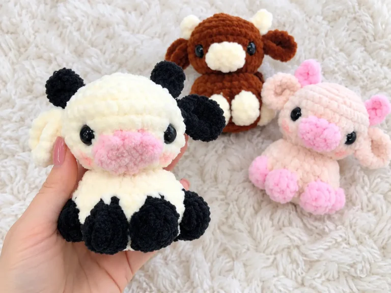 Hand holding a cream and black plush cow amigurumi toy with a pink snout on a white fluffy blanket.