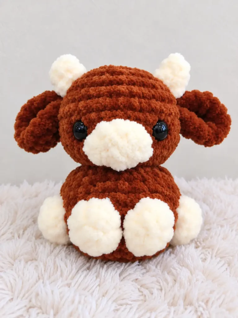 Brown and beige plush cow amigurumi toy with white horns sitting on a fuzzy white blanket.