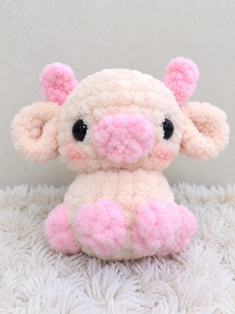 Light pink and cream plush cow amigurumi toy with a bright pink snout sitting on a white blanket.
