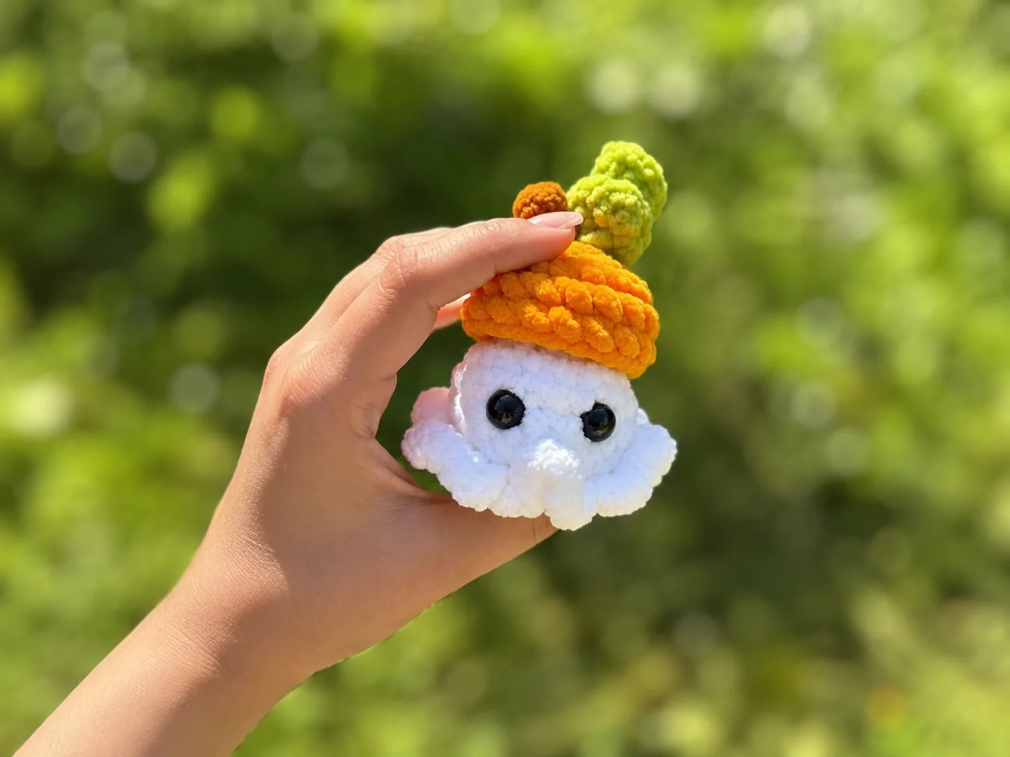 A white chenille crochet ghost with an orange pumpkin-style hat and green leaf sitting on a grey couch cushion.