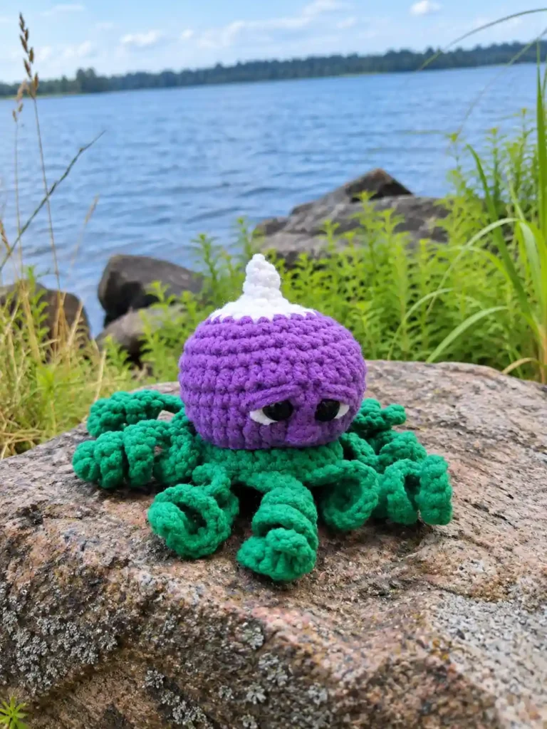 A purple radish octopus crochet pattern amigurumi with green tentacles sitting on a large rock by a blue lake.