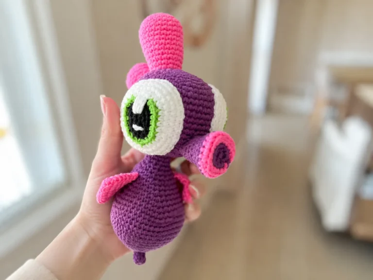 A deep purple crochet sea horse amigurumi with pink crests and fins, resting on a white blanket.