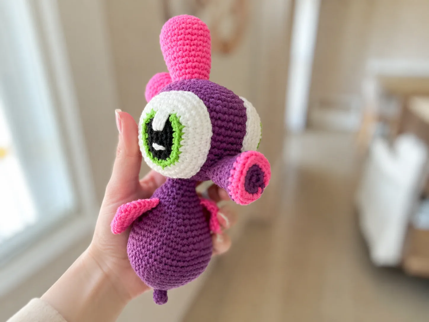 A deep purple crochet sea horse amigurumi with pink crests and fins, resting on a white blanket.